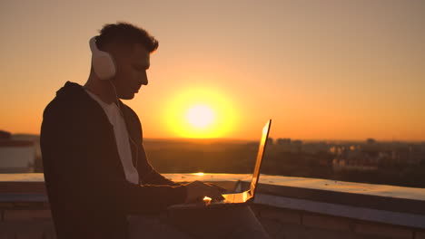 Ein-Mann-Mit-Kapuzenpullover-Sitzt-Auf-Dem-Dach-Und-Hört-Musik-Mit-Kopfhörern-Und-Tippt-Auf-Der-Tastatur-Eines-Laptops.-Laufen-Bei-Sonnenuntergang.-Freiberufler-Arbeitet-Bei-Sonnenuntergang-Mit-Einem-Laptop